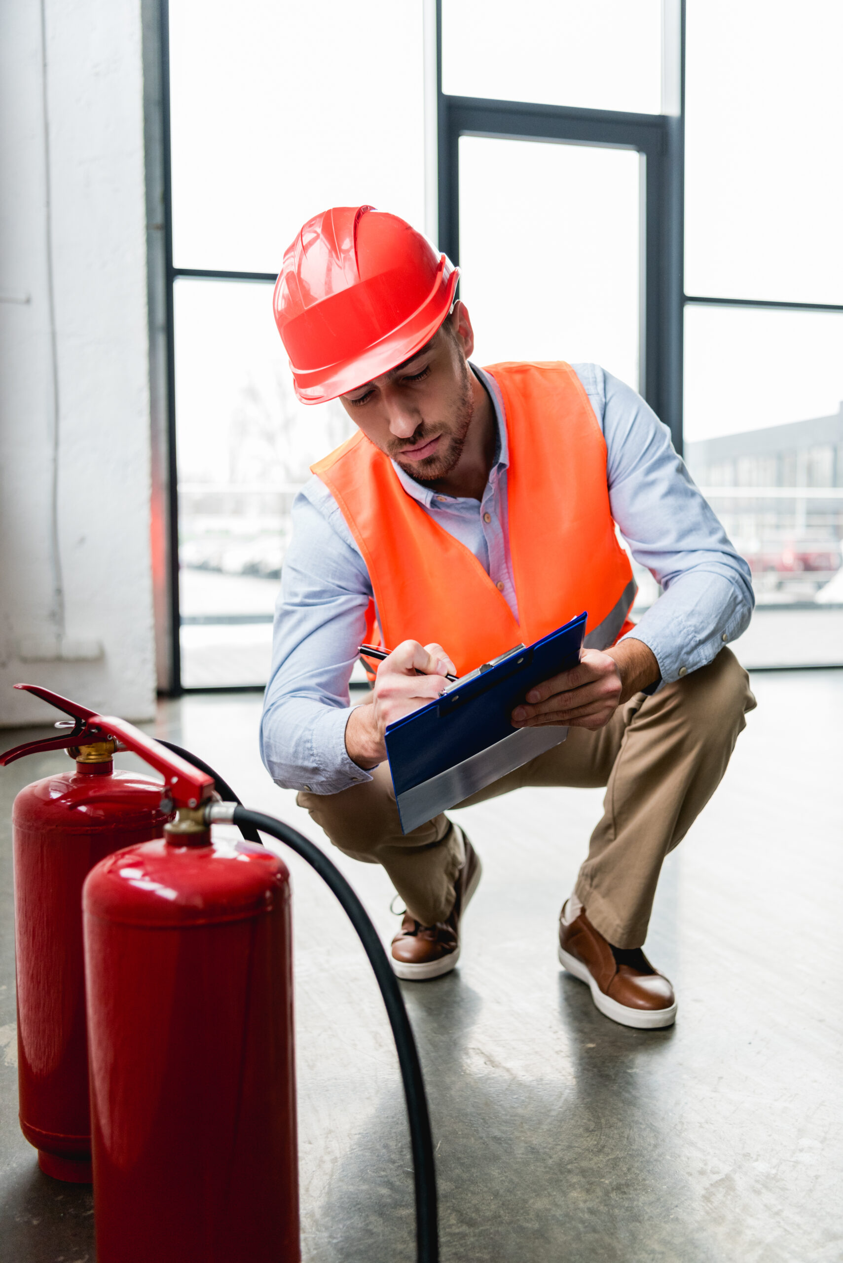 serious fireman in helmet writing on clipboard while checking extinguishers